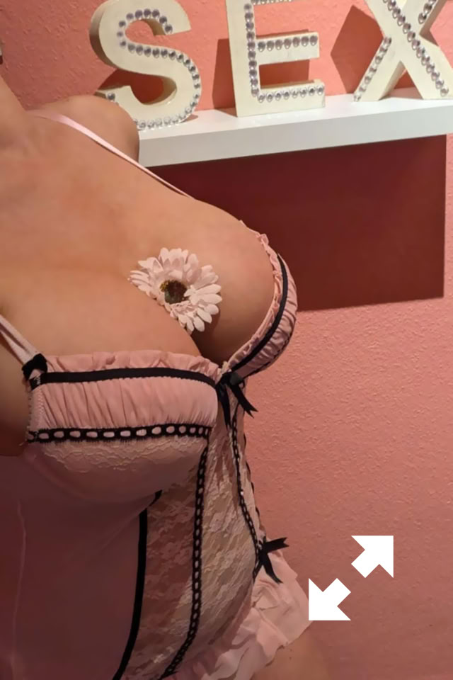 Celine in rosa Dessous mit schwarzen Details, eine Blume auf der Brust, vor einer rosa Wand mit dem Wort "SEX" in weißen Buchstaben.