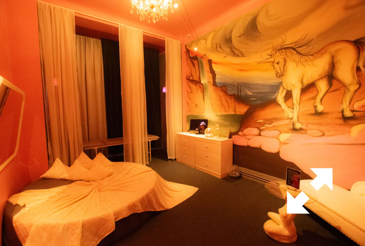 A room with pink walls, a bed with a draped cover, a mural of horses, a chandelier, and a dresser with a small television.