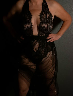 Jill wearing a sheer black lace outfit with hands on hips, standing against a dark background.