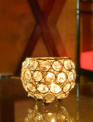 Ein dekoratives Teelicht aus Glas mit runden Kristallen auf einem Tisch, im Hintergrund ein Teil eines Bettes mit roten und schwarzen Kissen.