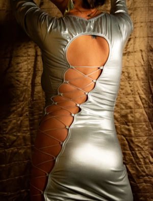 Nora wearing a shiny silver dress with a large cutout and lace-up detail on the back, standing against a textured brown background.