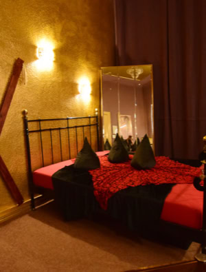 Zimmer 1 mit einem Metallbett, roten und schwarzen Bettdecken, einem großen Spiegel, braunen Vorhängen und einer Holzkonstruktion an der Wand.