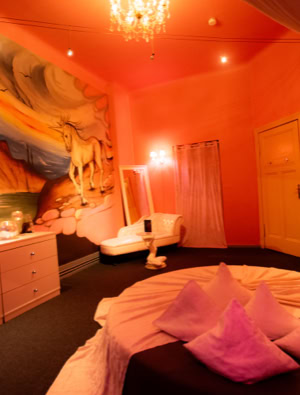 Zimmer 2 mit orangefarbener Beleuchtung, einem runden Bett mit Kissen, einer Kommode mit Fernseher, einem Wandgemälde und einem Kronleuchter.